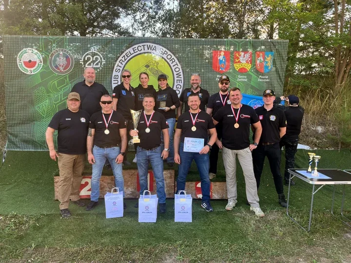 Grupa osób w czarnych koszulkach z medalami, pucharem i dyplomami pozuje na podium podczas zawodów strzeleckich w Zamościu.