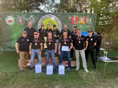 Grupa osób w czarnych koszulkach z medalami, pucharem i dyplomami pozuje na podium podczas zawodów strzeleckich w Zamościu.