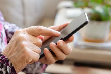Starsza osoba trzyma w dłoniach smartfon i dotyka ekranu palcem w wnętrzu pomieszczenia.